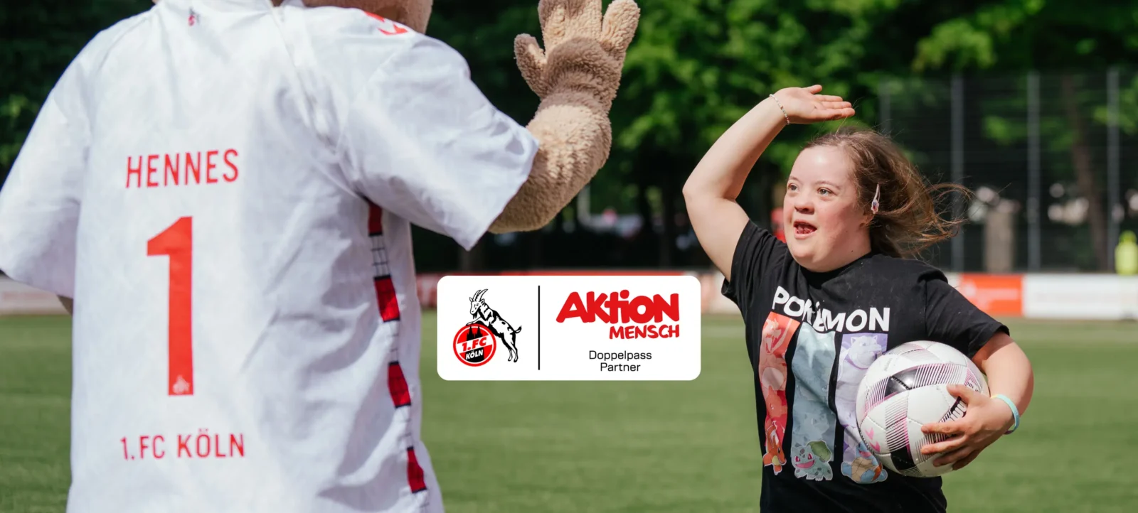Ein junges Mädchen mit Down-Syndrom spielt Fußball auf einem Rasenplatz. Sie trägt ein schwarzes T-Shirt mit einem Pokémon-Motiv und hält einen Fußball in den Händen. Neben ihr steht die Mascot "Hennes" des 1. FC Köln, ein braunes Stofftier, das ein weißes Trikot mit der Nummer 1 und dem Vereinslogo trägt. Im Vordergrund ist ein Schild mit dem Logo des 1. FC Köln und der Aufschrift "Aktion Mensch Doppelpass Partner" zu sehen.