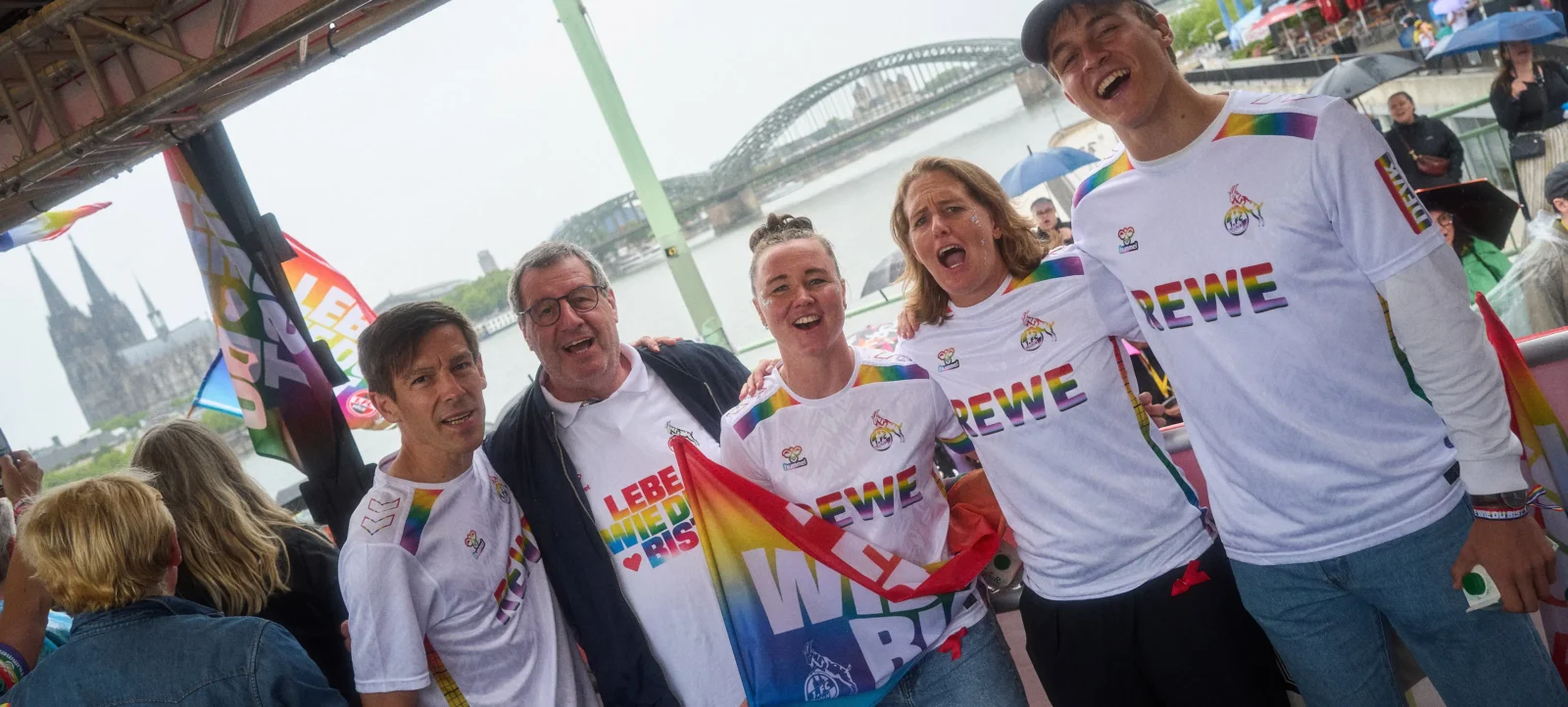 Eine Gruppe von Menschen steht auf einem Boot und lacht fröhlich. Sie tragen weiße T-Shirts mit dem Aufdruck "DEWE" und Regenbogen-Elementen. Eine Person hält eine Regenbogenfahne mit der Aufschrift "LEBE WIE DU BIST". Im Hintergrund ist die Kölner Brücke zu sehen.