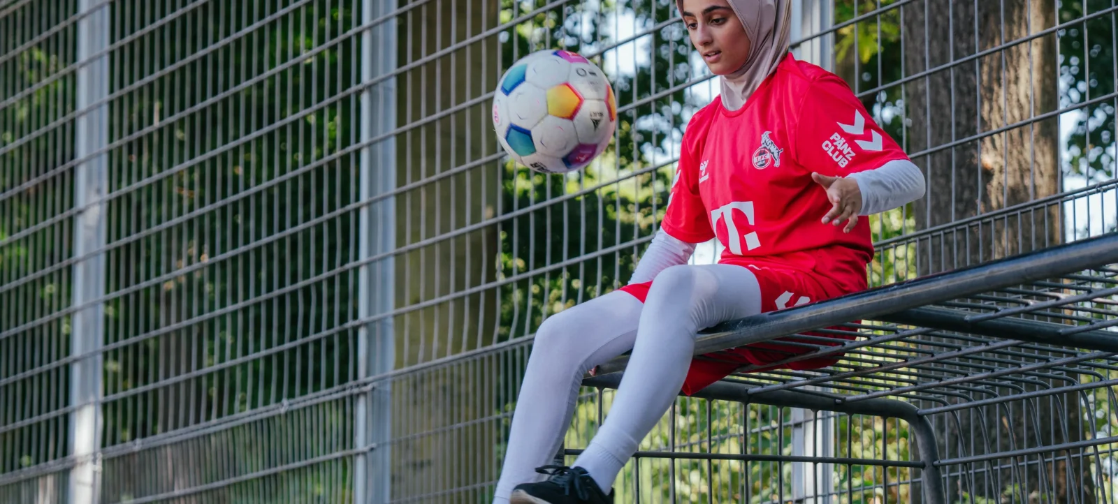Eine Person in roter Sportkleidung und einem grauen Kopftuch sitzt auf einem Metallzaun und hält einen bunten Fußball in der Hand. Im Hintergrund sind Bäume und ein Zaun zu sehen.