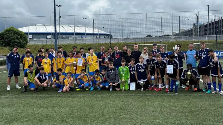Eine Gruppe von Kindern und Erwachsenen steht auf einem Fußballfeld für ein Gruppenfoto. Die Kinder tragen verschiedene Trikots in den Farben Blau, Gelb und Schwarz, einige halten Medaillen und Pokale. Die Erwachsenen, vermutlich Trainer oder Betreuer, stehen am Rand und tragen Trainingskleidung. Im Hintergrund ist ein Fußballstadion mit einem großen, kuppelartigen Dach zu sehen.