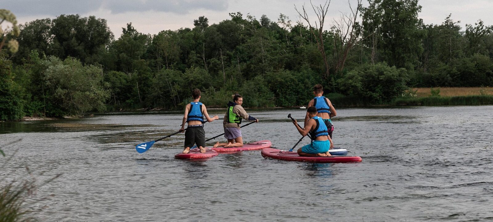 Vier Personen paddeln auf roten SUP-Brettern auf einem ruhigen Gewässer. Sie tragen Rettungswesten und bewegen sich in Richtung eines bewaldeten Ufers. Der Himmel ist leicht bewölkt, und die Umgebung wirkt friedlich und natürlich.