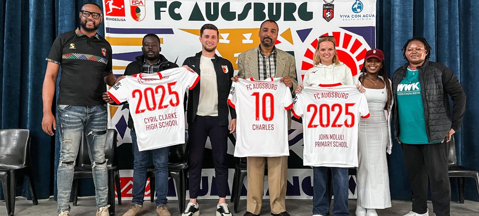 Eine Gruppe von sechs Personen steht vor einem Banner des FC Augsburg und hält Trikots mit den Aufschriften "FC Augsburg", "2025", "Cyril Clarke High School" und "John Moluli Primary School". Die Personen lächeln und posieren für ein Gruppenfoto.