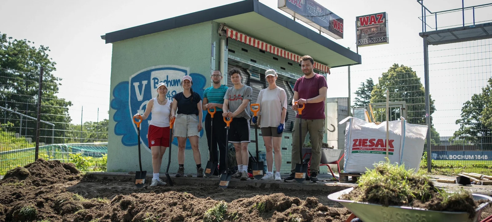 Eine Gruppe von sechs Personen steht vor einem Kiosk mit dem Logo des VFL Bochum und hält Schaufeln in den Händen. Sie scheinen an einem Gemeinschaftsprojekt oder einer Gartenarbeit teilzunehmen. Im Vordergrund sind Schubkarren mit Erde und Pflanzen zu sehen, und im Hintergrund befindet sich ein Zaun und ein Fußballfeld.