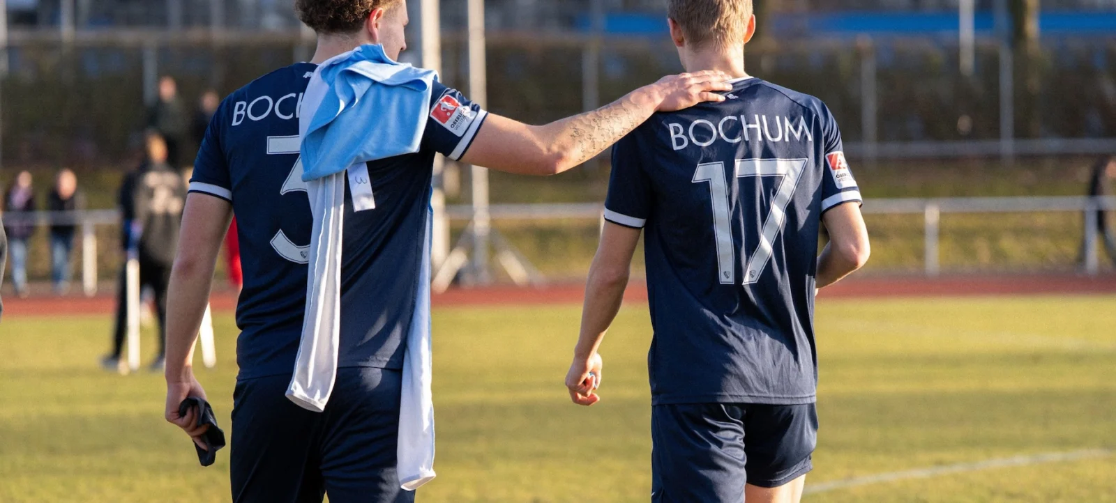 Zwei Fußballspieler des Vereins Bochum laufen gemeinsam vom Feld. Der Spieler mit der Nummer 17 hat den Arm um den Spieler mit der Nummer 3 gelegt, der ein blau-weißes Tuch über den Schultern trägt. Beide tragen dunkle Trikots mit dem Vereinsnamen "Bochum" auf dem Rücken. Im Hintergrund ist ein Fußballfeld mit einigen Personen zu sehen.