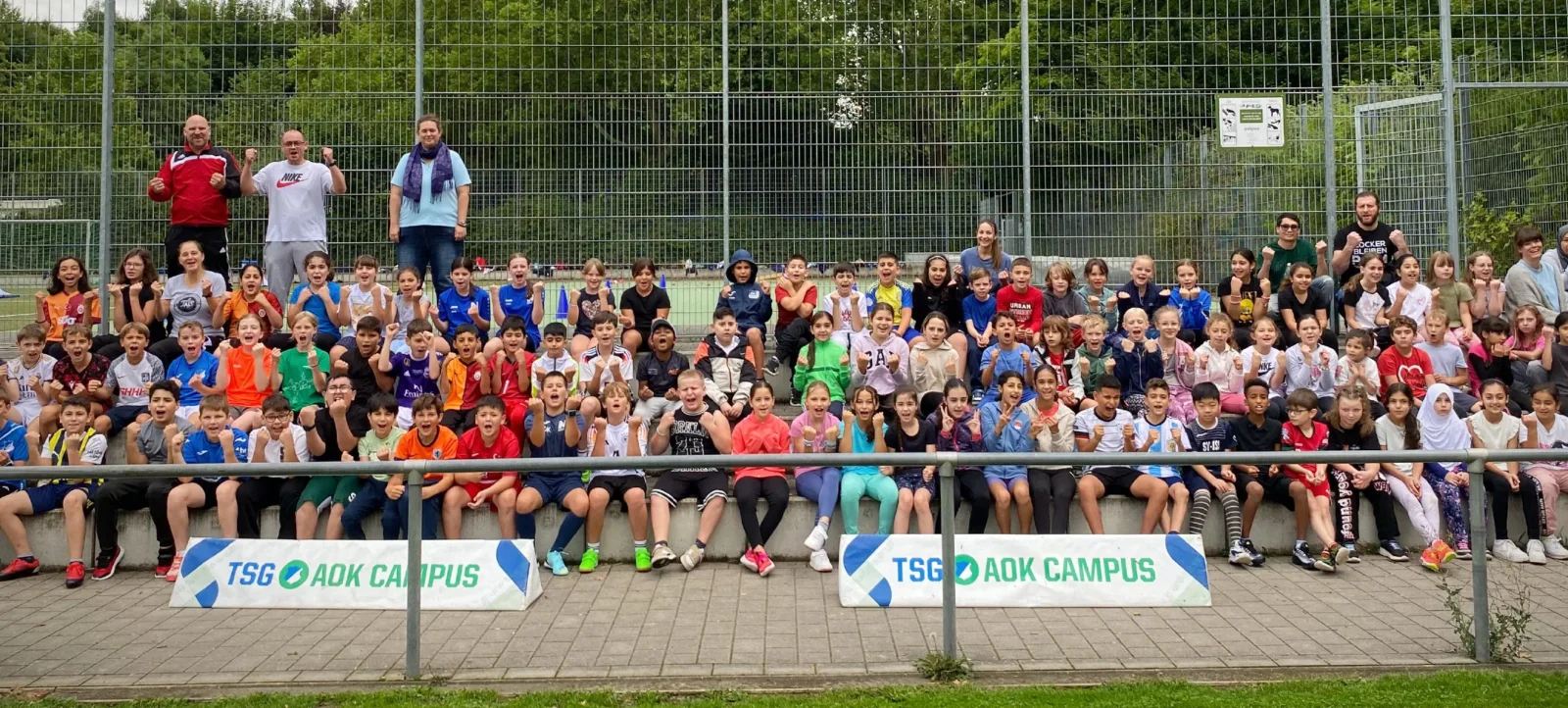 Eine große Gruppe von Kindern und Erwachsenen sitzt auf einer Treppe vor einem Zaun und posiert für ein Gruppenfoto. Die Kinder tragen bunte Kleidung und lächeln in die Kamera. Im Vordergrund sind zwei Schilder mit der Aufschrift "TSG AOK CAMPUS" zu sehen. Die Szene findet auf einem Sportgelände statt, umgeben von Grünflächen und Bäumen.