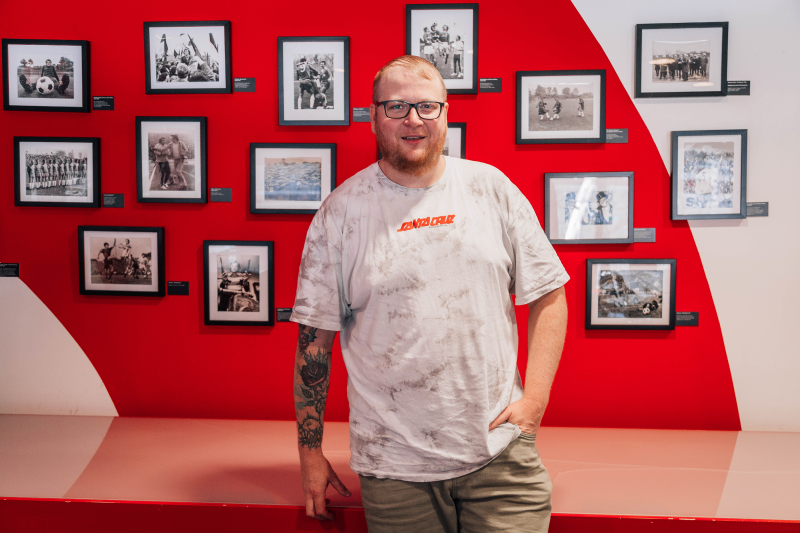 Ein Mann steht vor einer Wand mit historischen Schwarz-Weiß-Fotos, die Fußballszenen zeigen. Er trägt ein weißes T-Shirt mit einem Aufdruck und eine Brille. Die Fotos sind in schwarzen Rahmen angeordnet und zeigen verschiedene Momente aus der Fußballgeschichte.