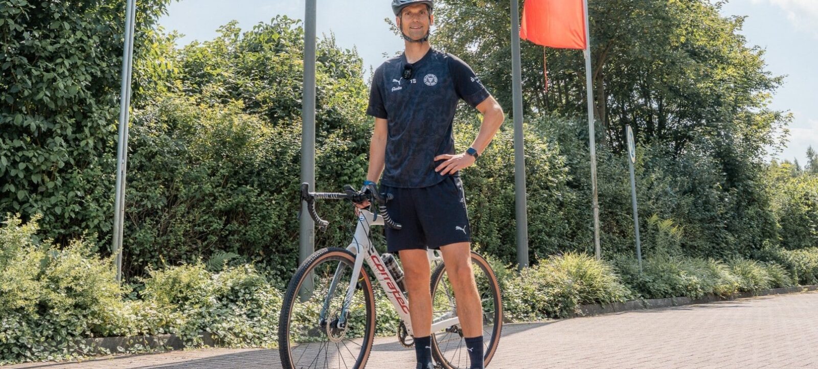 Ein Mann in Sportkleidung und Helm steht neben einem Rennrad auf einem gepflasterten Weg. Er trägt ein T-Shirt mit einem Logo und Shorts. Im Hintergrund sind grüne Büsche und Bäume sowie zwei Flaggen an Masten zu sehen.
