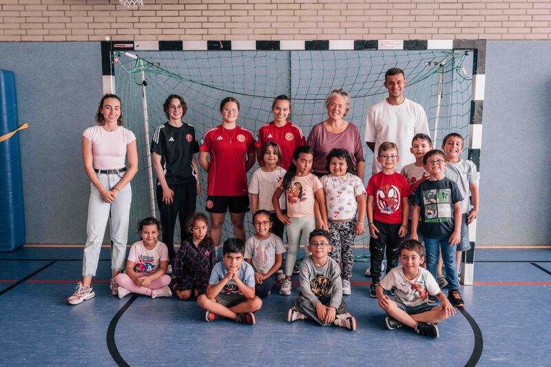 Eine Gruppe von Kindern und Erwachsenen posiert vor einem Tor in einer Sporthalle. Die Kinder sind in verschiedenen Altersgruppen und tragen unterschiedliche Kleidung, einige in Sportkleidung. Die Erwachsenen, darunter zwei Frauen und zwei Männer, stehen hinter den Kindern und lächeln in die Kamera. Die Sporthalle hat einen blauen Boden und eine weiße Torumrandung.