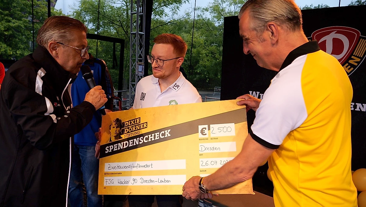 Drei Männer bei einer Spendenübergabe: Ein Mann in einem gelben Trikot überreicht einen großen Scheck über 2.500 Euro an einen Mann in weißem Trikot. Ein weiterer Mann mit Mikrofon steht daneben und kommentiert die Szene. Der Scheck ist an die FSG Häcker 90 Dresden-Leuben adressiert und trägt den Vermerk "Dixie Dörner Stiftungsscheck". Die Veranstaltung findet im Freien statt, im Hintergrund sind Bäume und eine Bühne zu sehen.