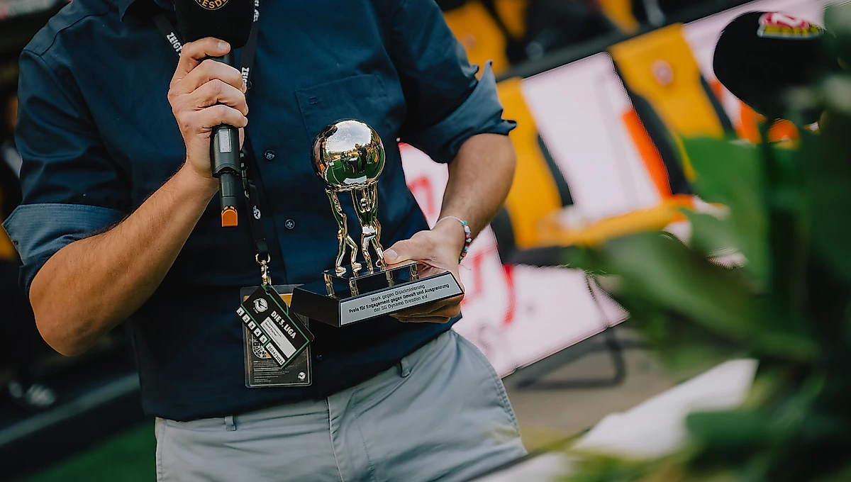 Ein Mann hält einen glänzenden Pokal in den Händen, während er ein Mikrofon in der anderen Hand hält. Im Hintergrund sind unscharf Flaggen und Menschen zu erkennen, was auf eine öffentliche Veranstaltung oder eine Preisverleihung hindeutet.
