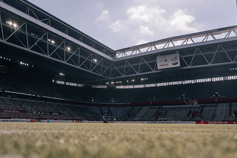 Ein weites Fußballstadion mit vielen Zuschauern auf den Rängen. Das Bild zeigt die Tribüne aus der Perspektive des Spielfelds. Die Tribünen sind mit Menschen gefüllt, die auf die Veranstaltung warten. Das Dach des Stadions ist offen und zeigt einen bewölkten Himmel.