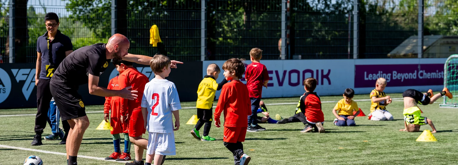 Ein Trainer erklärt einer Gruppe von Kindern auf einem Fußballfeld verschiedene Übungen. Die Kinder tragen verschiedene Trikots in den Farben Rot, Weiß, Gelb und Schwarz. Im Hintergrund ist eine Werbebande mit dem Slogan "Leading Beyond Chemistry" zu sehen.