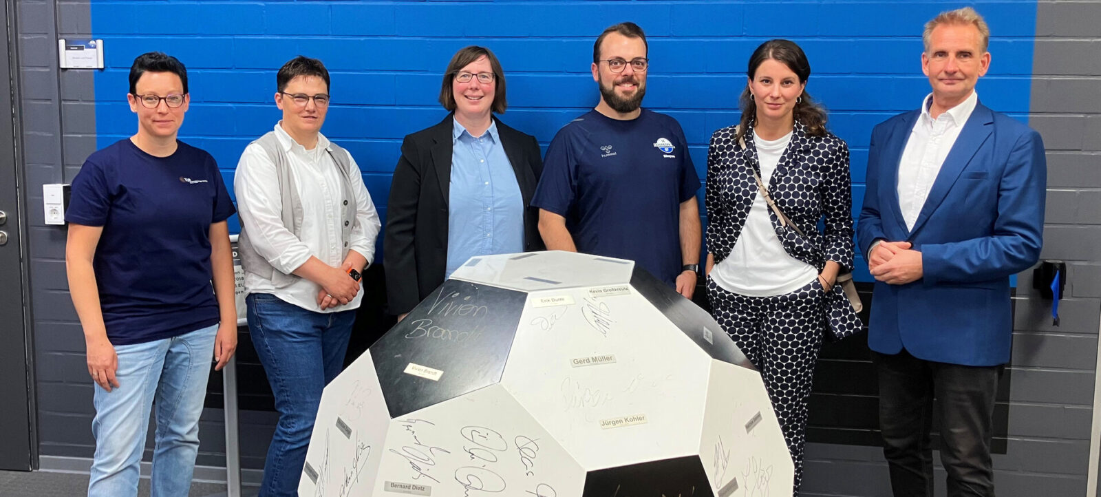 Eine Gruppe von sechs Personen steht vor einer blauen Wand und lächelt in die Kamera. Vor ihnen befindet sich ein großer, mit Autogrammen bedruckter Fußball. Die Personen sind unterschiedlich gekleidet, einige tragen Anzüge, andere legere Kleidung. Der Fußball trägt die Aufschrift "Vision Bayern" und zeigt zahlreiche Unterschriften bekannter Persönlichkeiten.