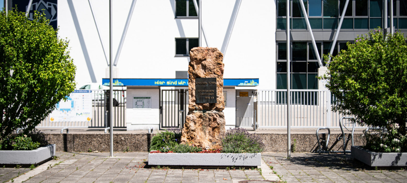 Ein Denkmal aus Stein mit einer Inschrift, umgeben von Blumen und Pflanzen, steht vor einem modernen Gebäude mit großen Fenstern und metallischen architektonischen Elementen. Im Hintergrund ist ein blaues Schild mit der Aufschrift "Hier sind wir" zu sehen.