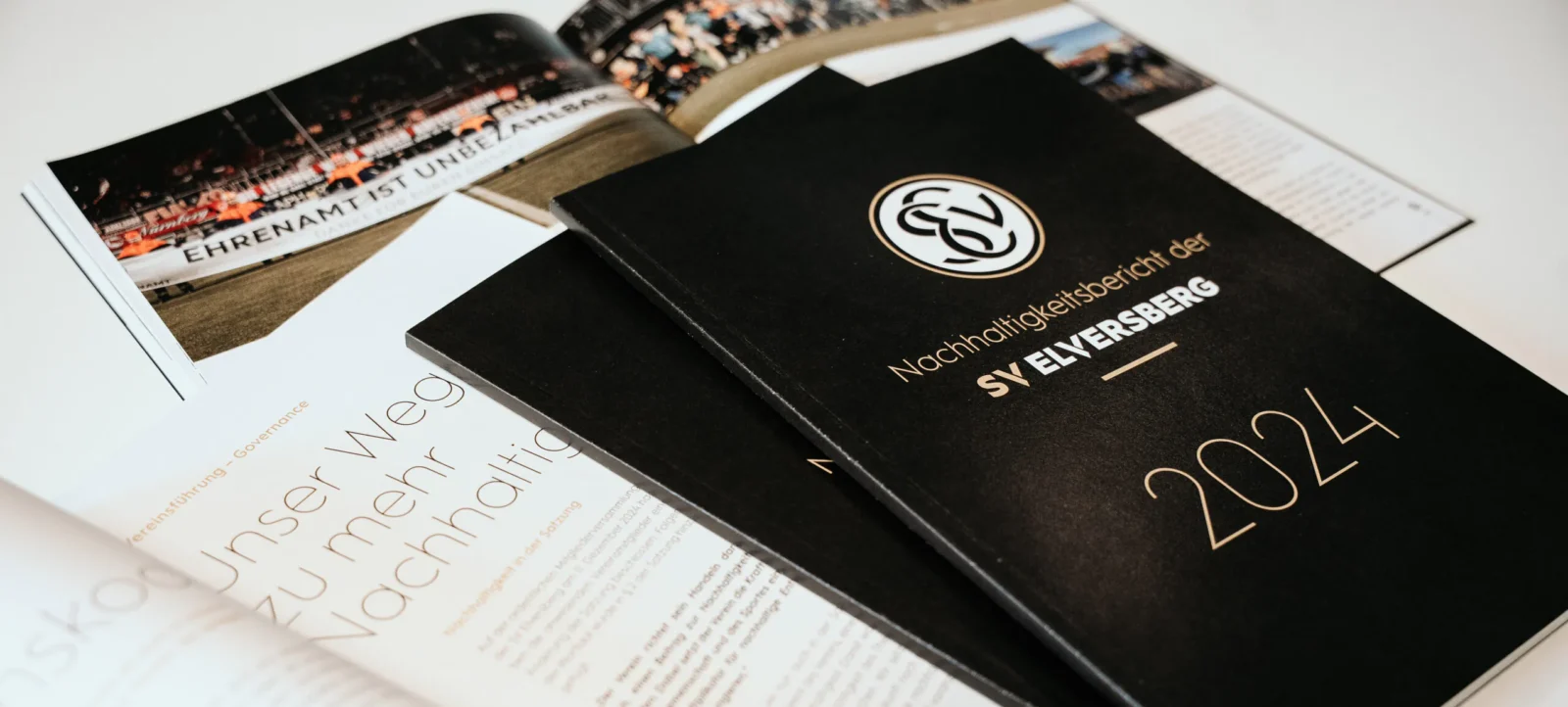 Ein aufgeschlagenes Nachhaltigkeitsbericht-Heft des SV Elversberg für das Jahr 2024 liegt auf einem Tisch. Das Heft zeigt auf der linken Seite einen Artikel mit dem Titel "Ehrenamt ist unbezahlbar" und auf der rechten Seite das Logo des SV Elversberg sowie den Text "Nachhaltigkeitsbericht der SV Elversberg 2024". Im Hintergrund sind Bilder von einem Fußballspiel zu sehen.