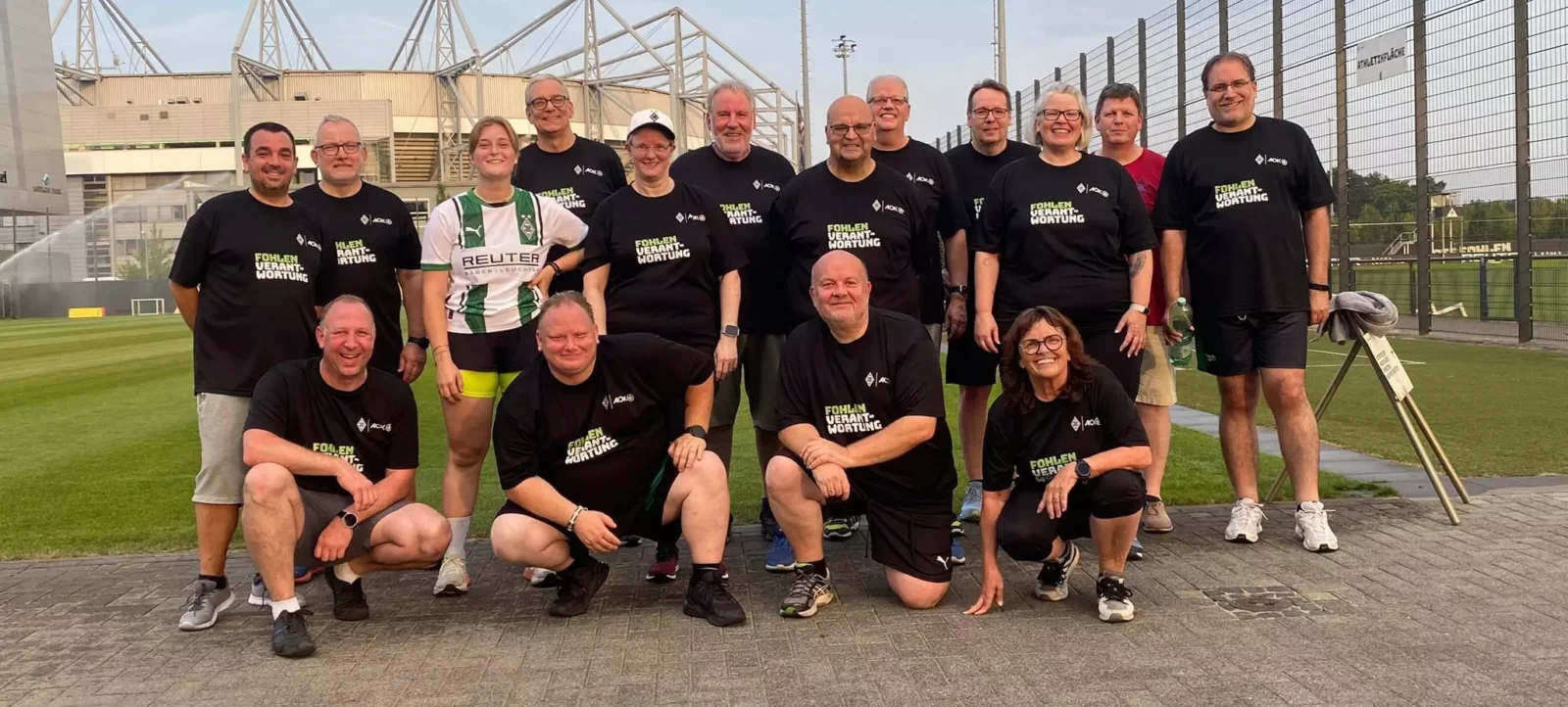 Eine Gruppe von Menschen steht und kniet vor einem Fußballstadion. Sie tragen schwarze T-Shirts mit dem Aufdruck "Fohlenverrückung" und einem Logo. Eine Person trägt ein grün-weißes Trikot mit dem Namen "Reuter". Im Hintergrund ist das Stadion mit seinen charakteristischen Stützen und einem Zaun zu sehen. Die Gruppe scheint für ein Gruppenfoto posiert zu haben.