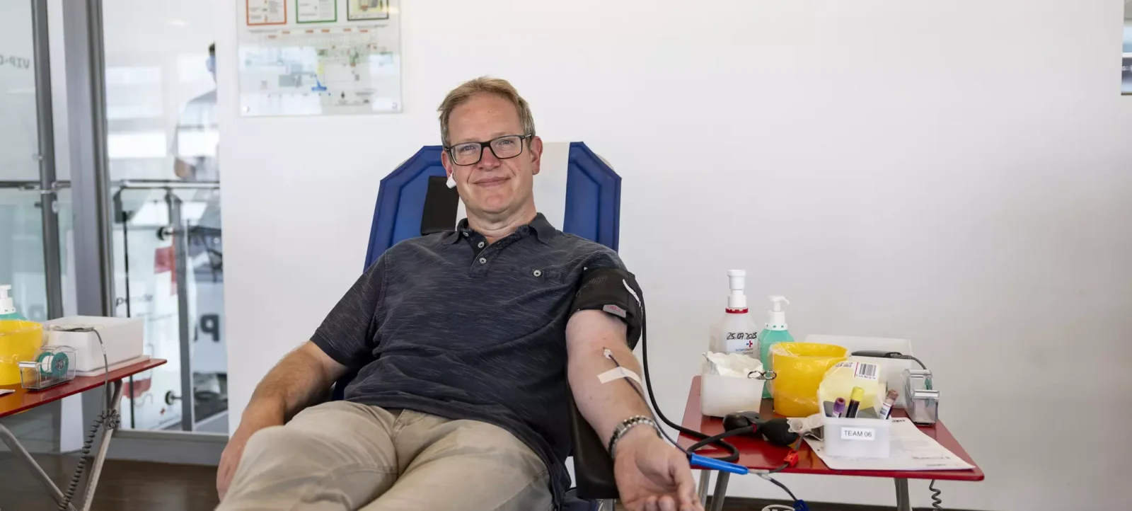 Ein Mann sitzt in einem bequemen Stuhl und spendet Blut. Er trägt ein schwarzes T-Shirt und eine Brille. Im Hintergrund ist ein Flucht- und Rettungsplan an der Wand zu sehen. Auf dem Tisch neben ihm befinden sich verschiedene Gegenstände wie Desinfektionsmittel, Handschuhe und ein Klemmbrett mit der Aufschrift "TEAM 06".
