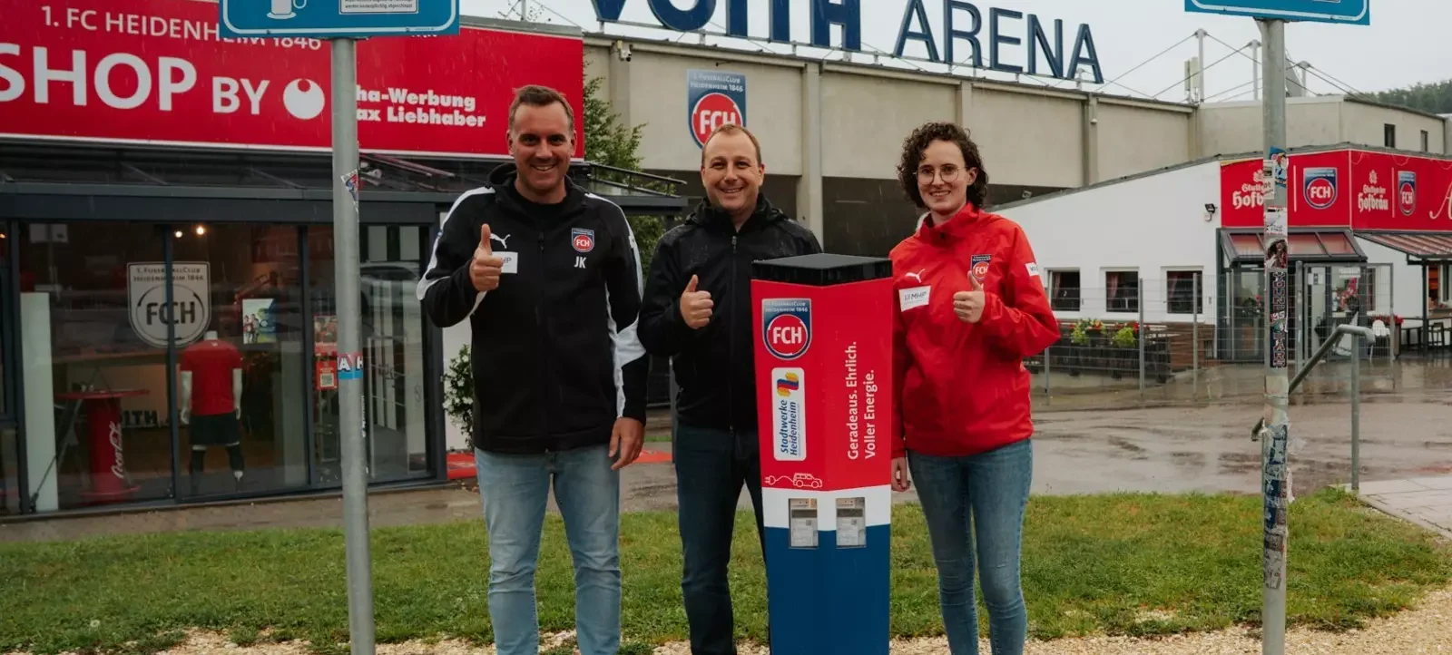 Drei Personen stehen vor einer Ladestation für Elektrofahrzeuge an der Voith Arena, dem Stadion des 1. FC Heidenheim. Sie tragen Jacken mit dem Logo des 1. FC Heidenheim und halten ihre Daumen hoch. Die Ladestation ist mit dem Logo des 1. FC Heidenheim und dem Slogan "Geradeaus. Ehrlich. Volle Energie." versehen. Im Hintergrund ist der Shop des 1. FC Heidenheim zu sehen.