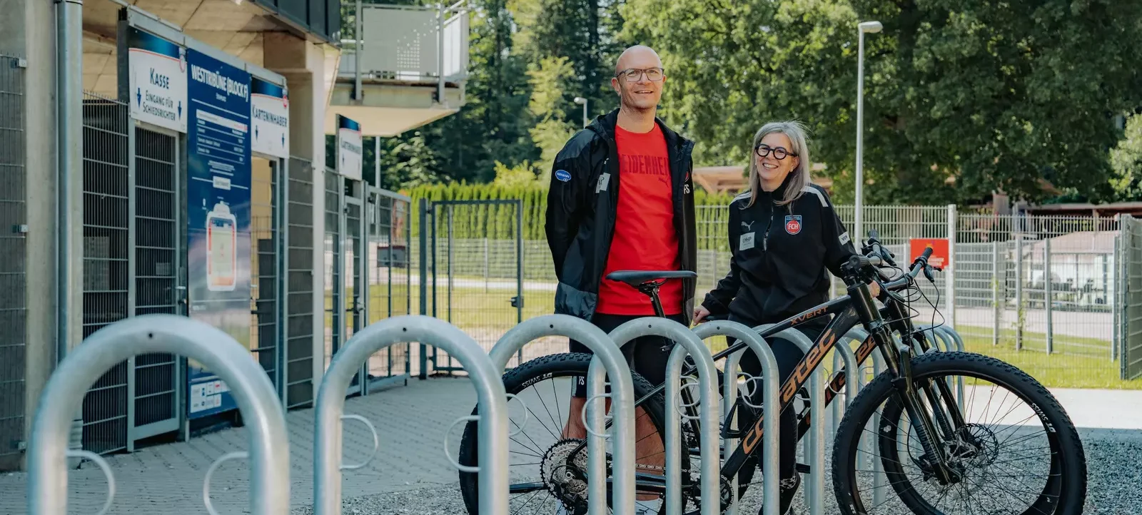Zwei Personen stehen vor einem Fahrradabstellplatz an einem Stadion. Die eine Person trägt eine schwarze Jacke über einem roten T-Shirt und die andere eine schwarze Jacke mit einem Vereinslogo. Sie lächeln in die Kamera. Im Hintergrund sind Schilder mit den Aufschriften "Kasse", "Eingang für Schiedsrichter" und "Westtribüne Block C" zu sehen.