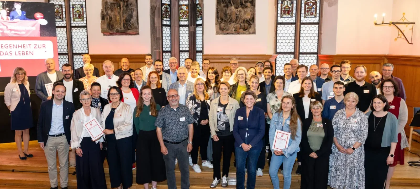 Eine Gruppe von Menschen steht in einer Kirche und lächelt in die Kamera. Die Gruppe besteht aus Erwachsenen verschiedener Altersgruppen und sie halten Urkunden in den Händen. Im Hintergrund sind bunte Bleiglasfenster und Wandgemälde zu sehen. Auf der linken Seite ist ein Teil eines Plakats mit der Aufschrift "EIGENHEIT ZUR DAS LEBEN" sichtbar. Die Atmosphäre wirkt feierlich und gemeinschaftlich.