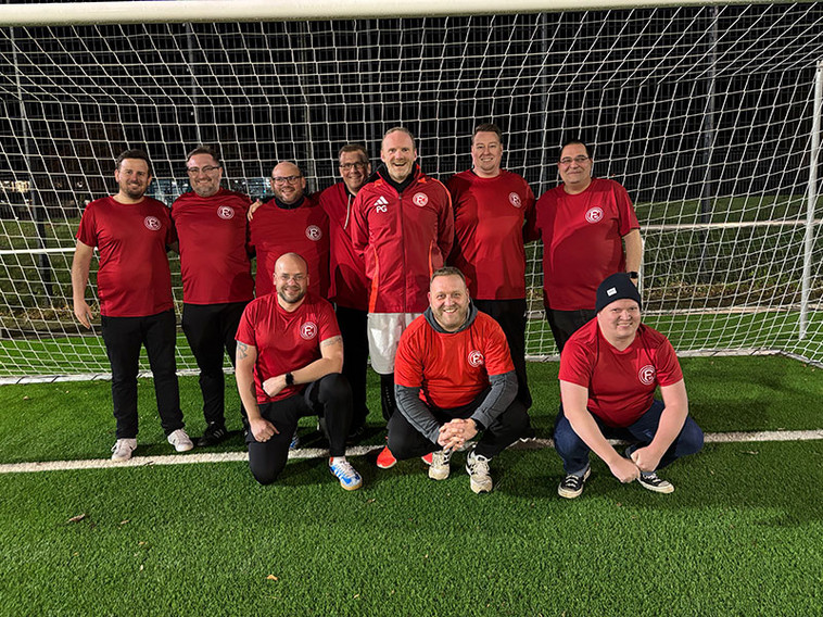 Eine Gruppe von Fußballfans in roten Trikots posiert vor einem Tor auf einem Rasenplatz. Die Fans stehen und knien in zwei Reihen, lächelnd und entspannt.
