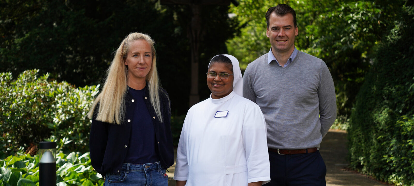 Drei Personen stehen im Freien vor einer grünen Hecke. Eine Frau mit blonden Haaren trägt eine schwarze Jacke und Jeans. Neben ihr steht eine Frau in weißer Krankenschwesterntracht mit einem Namensschild. Rechts steht ein Mann mit kurzen Haaren, der ein graues Pullover und eine dunkle Hose trägt. Alle lächeln in die Kamera.