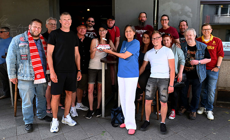 Eine Gruppe von Menschen steht vor einem Gebäude und lächelt in die Kamera. Eine Frau in der Mitte hält einen Scheck in den Händen und überreicht ihn einer anderen Frau, die ein blaues T-Shirt und weiße Hosen trägt. Die Gruppe besteht aus Männern und Frauen unterschiedlichen Alters, einige tragen Sportbekleidung und Schals mit Vereinslogos. Die Atmosphäre wirkt fröhlich und gesellig, möglicherweise handelt es sich um eine Spendenübergabe oder ein gemeinsames Projekt.