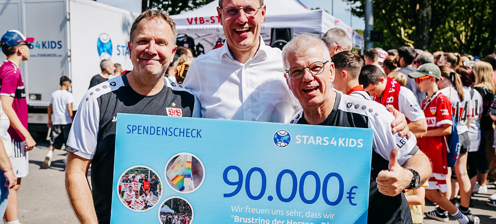 Drei Männer halten einen großen Scheck über 90.000 Euro für das Projekt "Stars4Kids" der VFB-Stiftung. Sie lächeln und stehen vor einer Gruppe von Menschen, die an einem Event teilnehmen. Im Hintergrund sind ein weißer Transporter und ein Zelt mit dem Logo des VfB zu sehen.