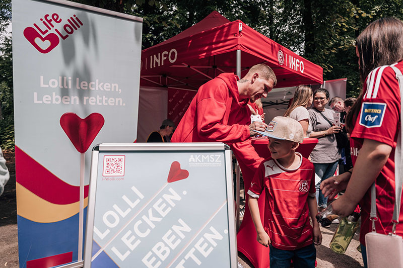 Ein Mann in rotem T-Shirt überreicht einem Kind in rotem T-Shirt und weißer Mütze einen Lolli an einem Infostand mit dem Logo "Life Lolli" und dem Slogan "Lolli lecken, Leben retten". Im Hintergrund stehen weitere Personen, darunter eine in rotem Trikot mit blauem Vereinslogo.