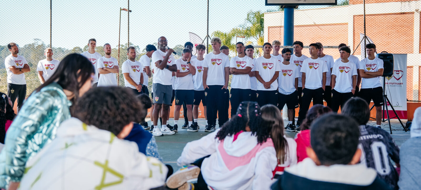 Eine Gruppe von jungen Männern in weißen T-Shirts mit einem roten und gelben Logo steht auf einem Sportplatz. Sie scheinen Teil eines Teams zu sein und hören aufmerksam einem Mann zu, der mit einem Mikrofon spricht. Im Vordergrund sitzen mehrere Kinder auf dem Boden und schauen zu der Gruppe hinauf. Im Hintergrund ist ein Basketballkorb zu sehen.