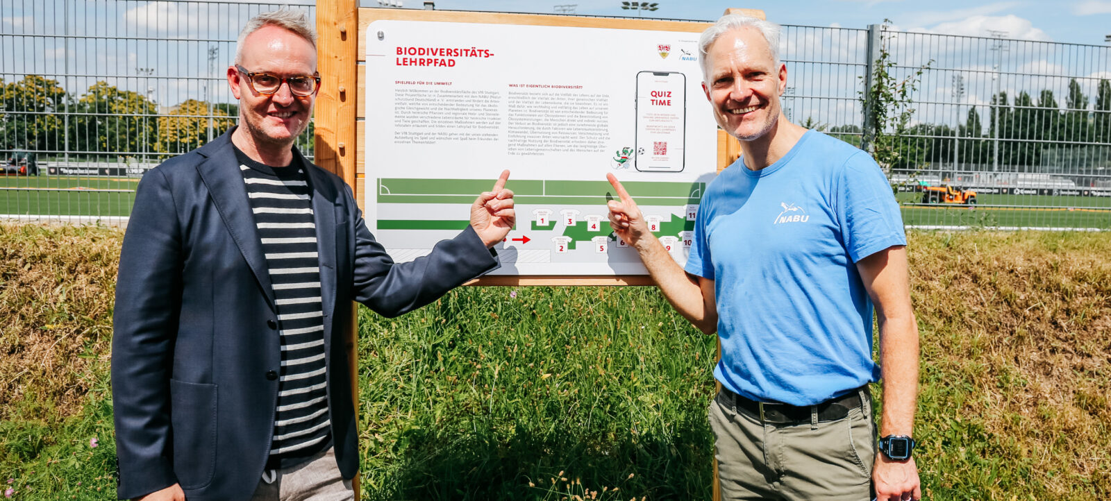 Zwei Männer stehen vor einem Schild mit der Aufschrift "Biodiversitäts-Lehrpfad" und zeigen auf ein Quiz-Diagramm. Der Mann links trägt eine Brille, einen dunklen Blazer und ein gestreiftes Hemd. Der Mann rechts trägt ein blaues T-Shirt mit dem Logo "NABU" und hält eine blaue Kappe in der Hand. Im Hintergrund ist ein Fußballfeld mit Zaun und Bäumen zu sehen.