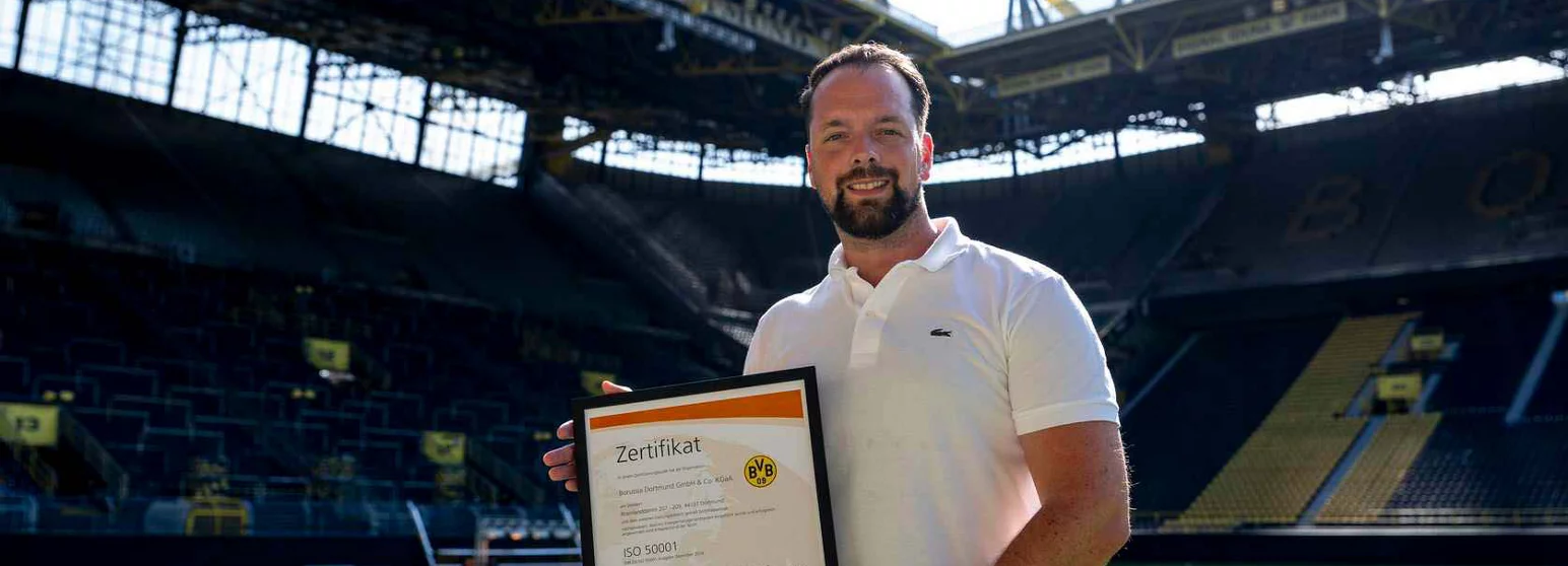 Ein Mann in weißem Polo-Shirt hält ein Zertifikat in den Händen und steht in einem großen, teilweise beleuchteten Stadion. Das Zertifikat trägt den Titel "Zertifikat" und scheint mit dem Logo "BVB" und der ISO-Norm 50001 versehen zu sein.