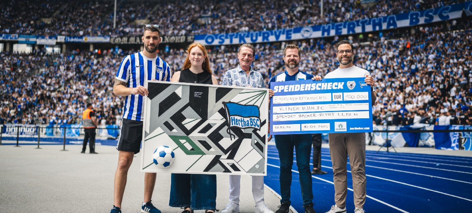 Vier Personen stehen auf einem Fußballfeld und halten einen großen Scheck und ein Kunstwerk. Der Scheck zeigt eine Spende von 100.000 Euro von "Becher Rückgabe" an die "Kinderhilfe" des Hertha BSC. Die Personen stehen vor einem vollen Stadion mit vielen Zuschauern.