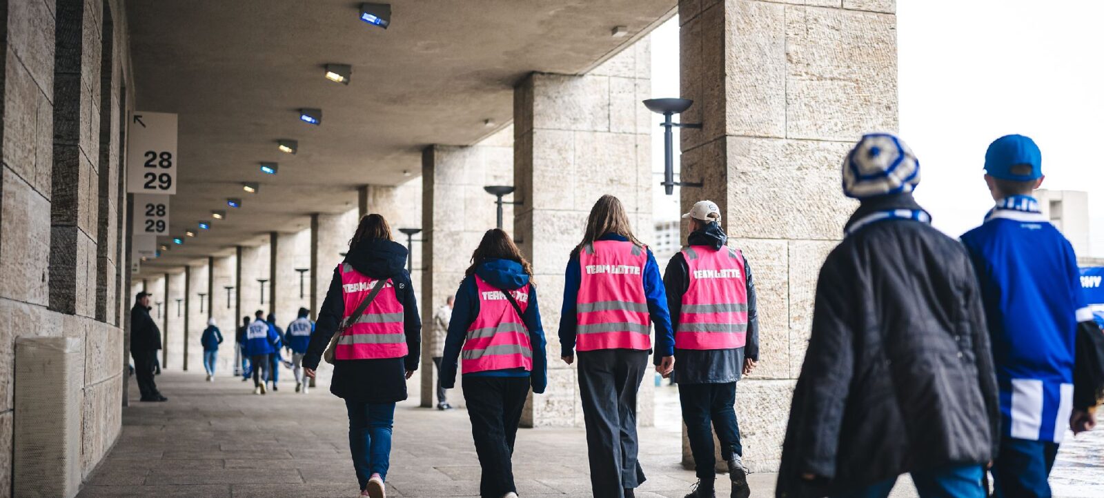 Eine Gruppe von Menschen, darunter mehrere Personen in pinken Westen mit der Aufschrift "Team Lotte", gehen durch einen überdachten Bereich mit Betonpfeilern. Die Szene scheint sich in einem Stadion oder einer großen Veranstaltungshalle zu befinden, wie die Schilder mit den Zahlen 28 und 29 an der Wand andeuten. Die Personen sind von hinten zu sehen und scheinen sich auf eine Veranstaltung oder ein Event vorzubereiten.