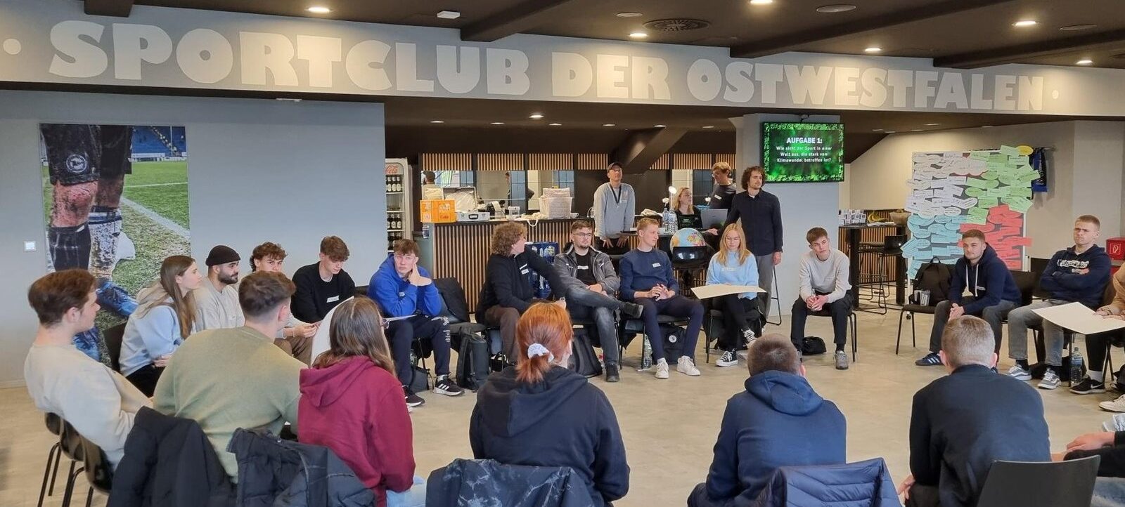 Eine Gruppe von Menschen sitzt in einem Kreis in einem Raum mit dem Schriftzug "SPORTCLUB DER OSTWESTFALEN" an der Wand. Die Teilnehmer scheinen an einer Diskussion oder einem Workshop teilzunehmen. Im Hintergrund sind Bildschirme und eine Theke mit Getränken und Snacks zu sehen.