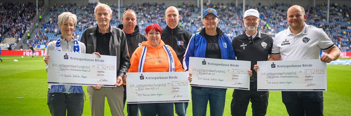 Eine Gruppe von sieben Personen steht auf einem Fußballfeld und hält Schecks der Kreissparkasse Börde in Höhe von jeweils 42.112,25 Euro. Die Schecks sind für den FCM Fanklub Sektion Angern bestimmt. Die Personen tragen verschiedene Kleidungsstücke, darunter Jacken und T-Shirts mit Vereinslogos. Im Hintergrund sind Zuschauer auf den Tribünen zu sehen.