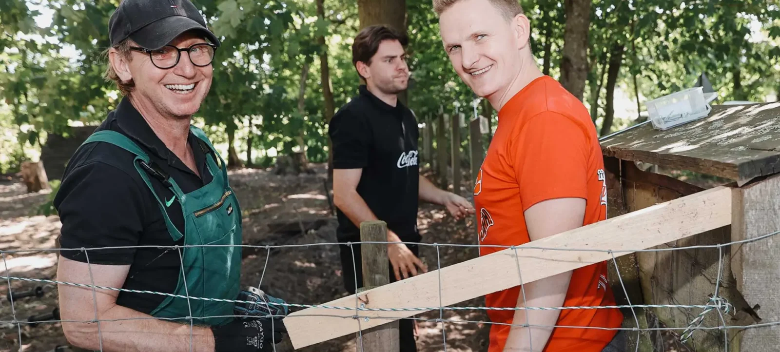 Drei Personen arbeiten auf einem Bauernhof. Zwei von ihnen, ein älterer Mann in grüner Latzhose und schwarzem Shirt sowie ein jüngerer Mann in orangefarbenem T-Shirt, lächeln und scheinen sich zu unterhalten. Sie stehen vor einem Holzzaun, der gerade repariert wird. Im Hintergrund ist ein weiterer Mann in schwarzem Shirt zu sehen, der ebenfalls am Hof arbeitet. Die Szene spielt in einem Waldgebiet mit viel Grün.