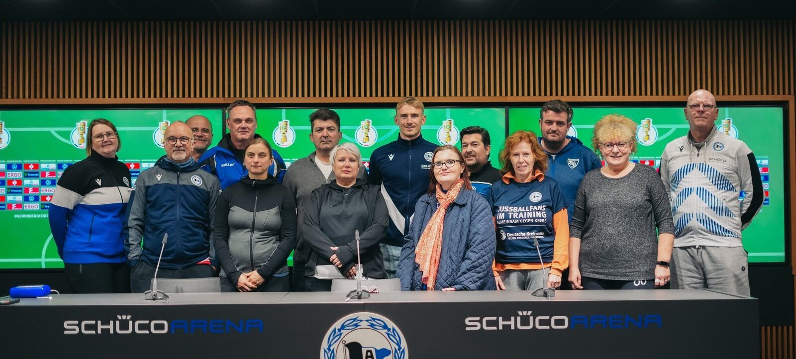 Eine Gruppe von Menschen steht hinter einem Podium in der SchücoArena. Sie tragen verschiedene Kleidungsstücke, darunter Jacken und T-Shirts, einige mit Logos von Vereinen oder Organisationen. Im Hintergrund ist eine große grüne Wand mit Logos und einem Tabellenbild zu sehen. Das Podium trägt das Logo des Vereins mit einem schwarzen und weißen Wappen und dem Schriftzug "SchücoArena". Die Atmosphäre wirkt formell, möglicherweise handelt es sich um eine Pressekonferenz oder eine offizielle Veranstaltung.