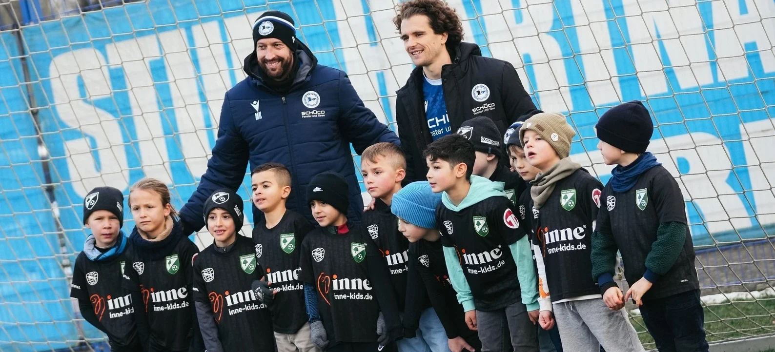 Zwei Erwachsene und eine Gruppe von Kindern stehen vor einem Fußballtor. Die Kinder tragen schwarze Jacken mit dem Aufdruck "Minne" und einem Logo. Die Erwachsenen lächeln und haben ihre Arme um die Kinder gelegt. Der Hintergrund zeigt ein blau-weißes Muster, möglicherweise Teil eines Stadions.
