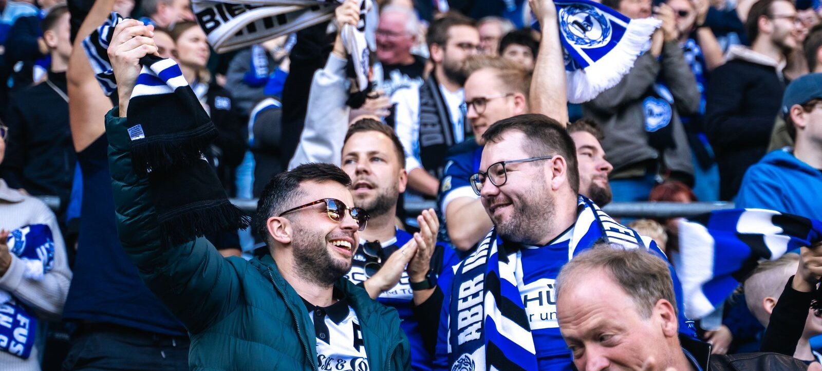 Eine Gruppe von Fußballfans im Stadion, die begeistert und freudig sind. Sie tragen Schals und Kleidung in den Vereinsfarben Blau und Weiß. Einige Fans halten Schals hoch und klatschen, während sie sich über ein Ereignis auf dem Spielfeld freuen.