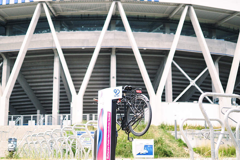 Ein Fahrrad steht an einem Fahrradverleihsystem vor einem modernen Stadion. Das Verleihsystem ist mit einem Schild versehen, das das Logo und den Namen des Systems zeigt. Im Hintergrund sind weitere Fahrradständer und ein Teil des Stadions mit seiner charakteristischen Architektur zu sehen.