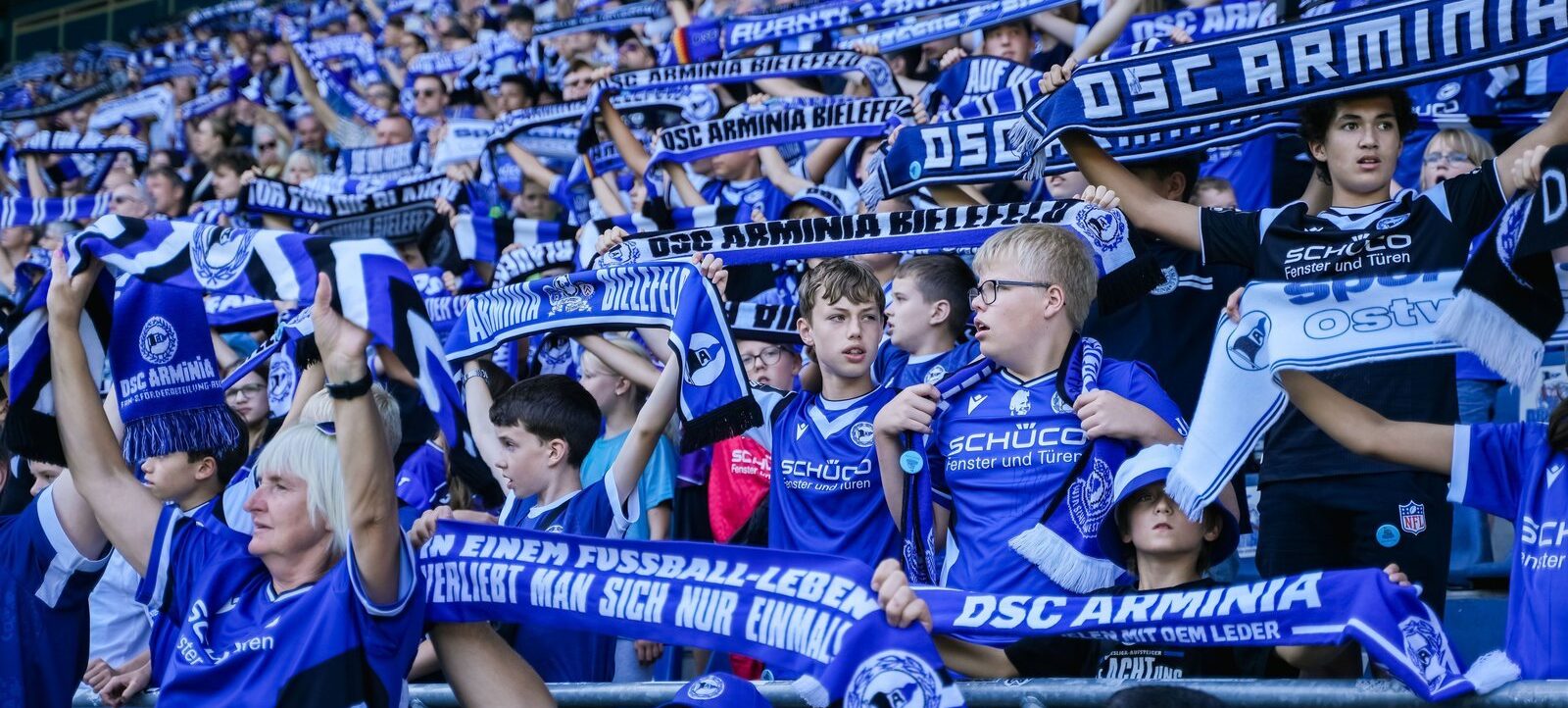 Eine Gruppe von Fans des DSC Arminia Bielefeld steht in den blauen und weißen Farben des Vereins in den Rängen eines Stadions. Sie halten Schals und Banner mit dem Vereinsnamen und Slogans hoch, während sie ein Fußballspiel unterstützen.