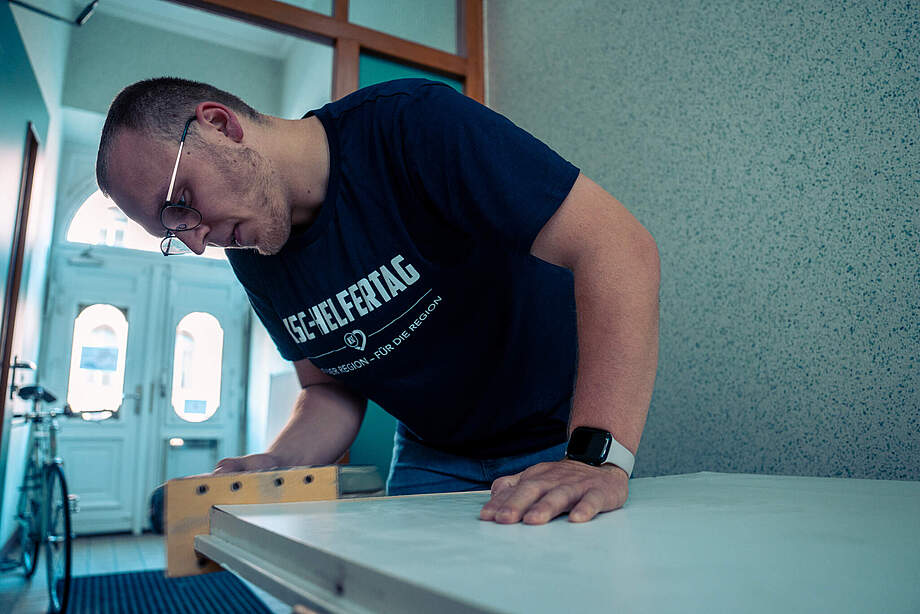 Ein Mann in einem T-Shirt mit der Aufschrift "1. FC Heidenheim 1846 Helfertag" arbeitet konzentriert an einer Werkbank. Er trägt eine Brille und eine Smartwatch und scheint mit einem Werkzeug an einem grauen Material zu arbeiten. Im Hintergrund sind ein Fahrrad und eine Tür mit runden Fenstern zu sehen.
