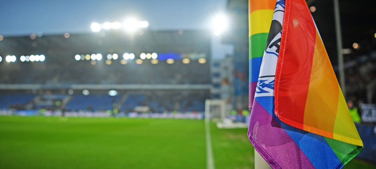 Ein Regenbogenfähnchen weht im Wind vor einem Fußballstadion. Im Hintergrund sind die beleuchteten Flutlichtmasten und der Rasenplatz zu sehen.
