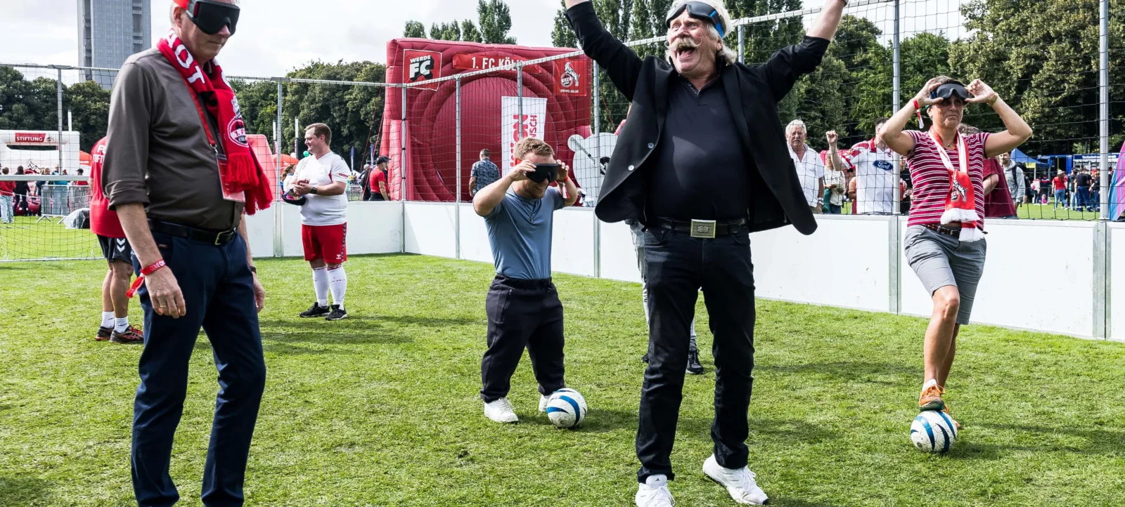 Eine Gruppe von Menschen spielt blind Fußball auf einem Rasenfeld. Die Spieler tragen Augenbinden und werden von Begleitpersonen unterstützt. Im Hintergrund ist ein Tor mit Werbung zu sehen.