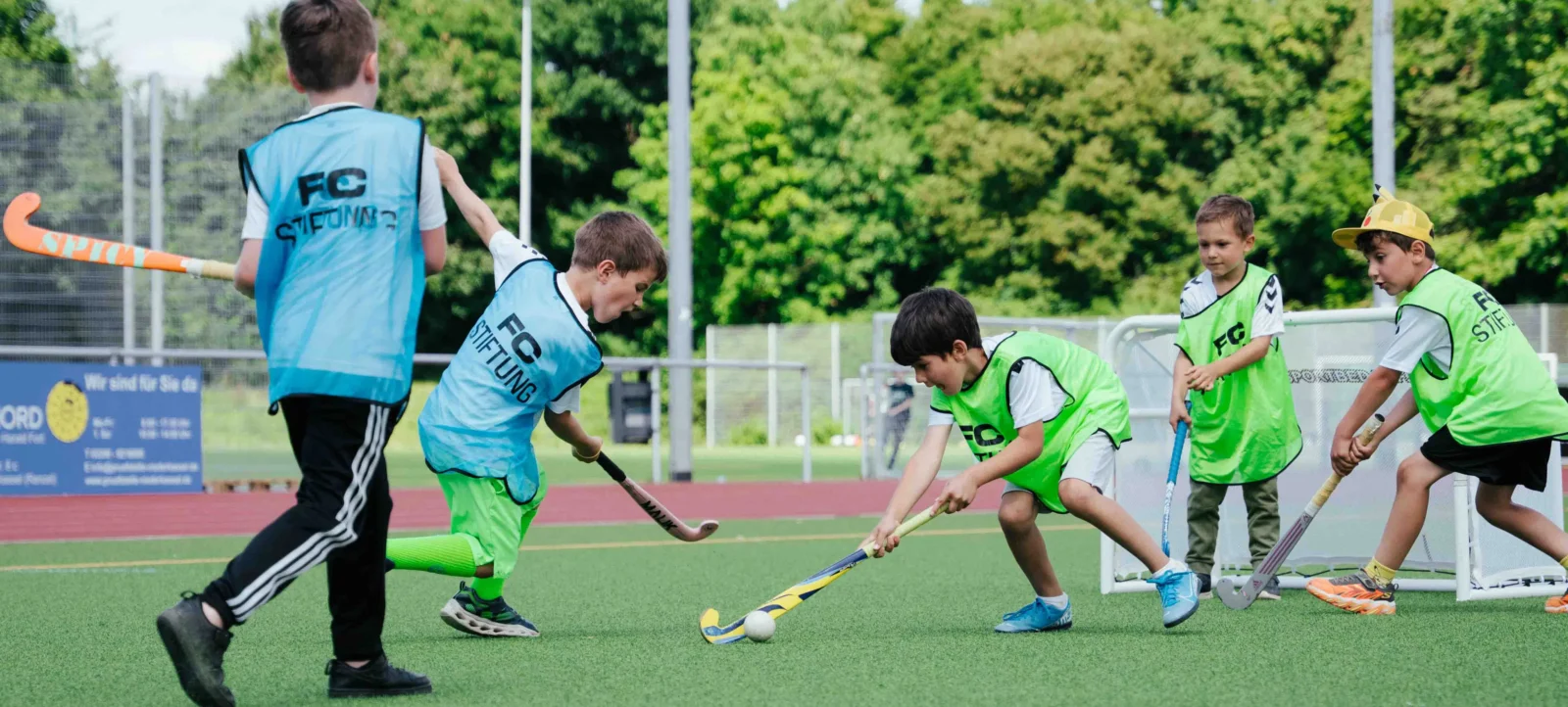 Kinder spielen Hockey auf einem Kunstrasenfeld. Sie tragen verschiedene farbige Trikots mit der Aufschrift 'FC STIFTUNG' und sind konzentriert auf den Ball gerichtet. Im Hintergrund sind grüne Bäume und ein Zaun zu sehen.