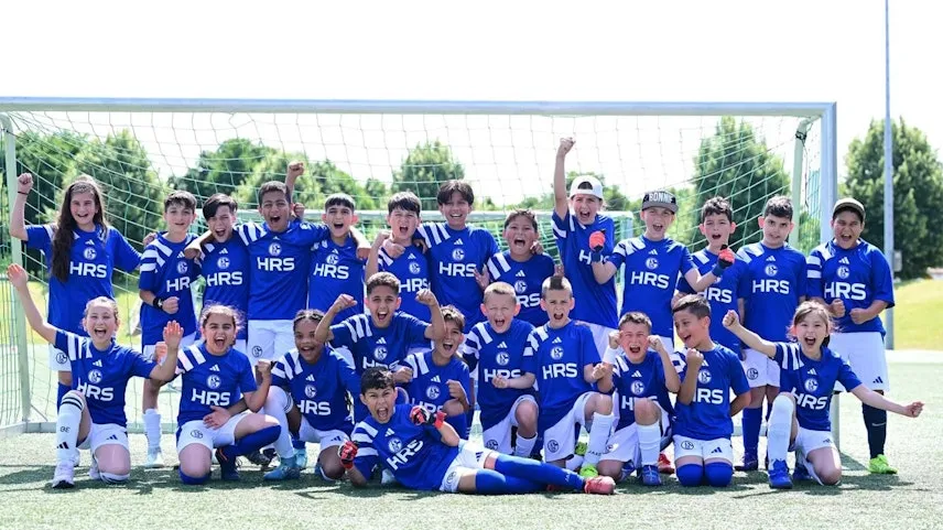 Eine Gruppe junger Fußballspieler in blauen Trikots mit dem Aufdruck "HRS" und "Adidas" posieren vor einem Tor für ein Gruppenfoto. Die Kinder strahlen und zeigen verschiedene Gesten der Freude und des Teamgeistes. Sie befinden sich auf einem Fußballfeld mit grünem Rasen und Bäumen im Hintergrund.