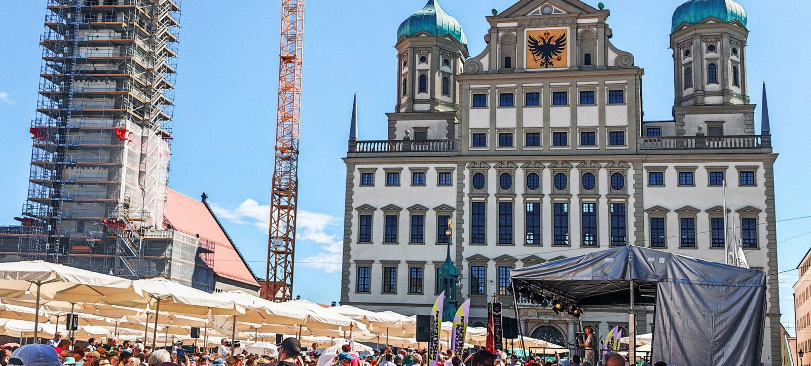 Ein belebter Platz vor einem historischen Gebäude mit zwei Türmen und einem Adler-Emblem. Im Hintergrund sind Baugerüste und ein Kran zu sehen, was auf laufende Renovierungsarbeiten hinweist. Menschenmengen genießen eine Veranstaltung unter großen Zelten, die mit Fahnen geschmückt sind.