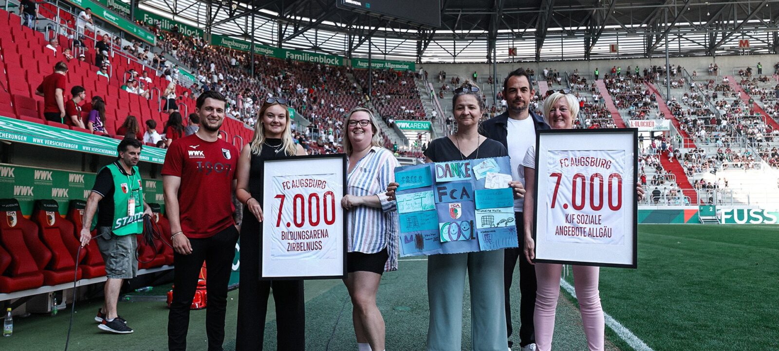 Eine Gruppe von fünf Personen steht auf dem Spielfeld eines Fußballstadions und hält Schilder hoch. Die Schilder zeigen die Aufschrift "FC Augsburg 7.000" und "KJF Soziale Angebote Allgäu". Die Personen lächeln und posieren für das Foto. Im Hintergrund sind Zuschauer auf den Tribünen zu sehen, und das Spiel steht 0:1. Die Atmosphäre wirkt feierlich und dankbar.