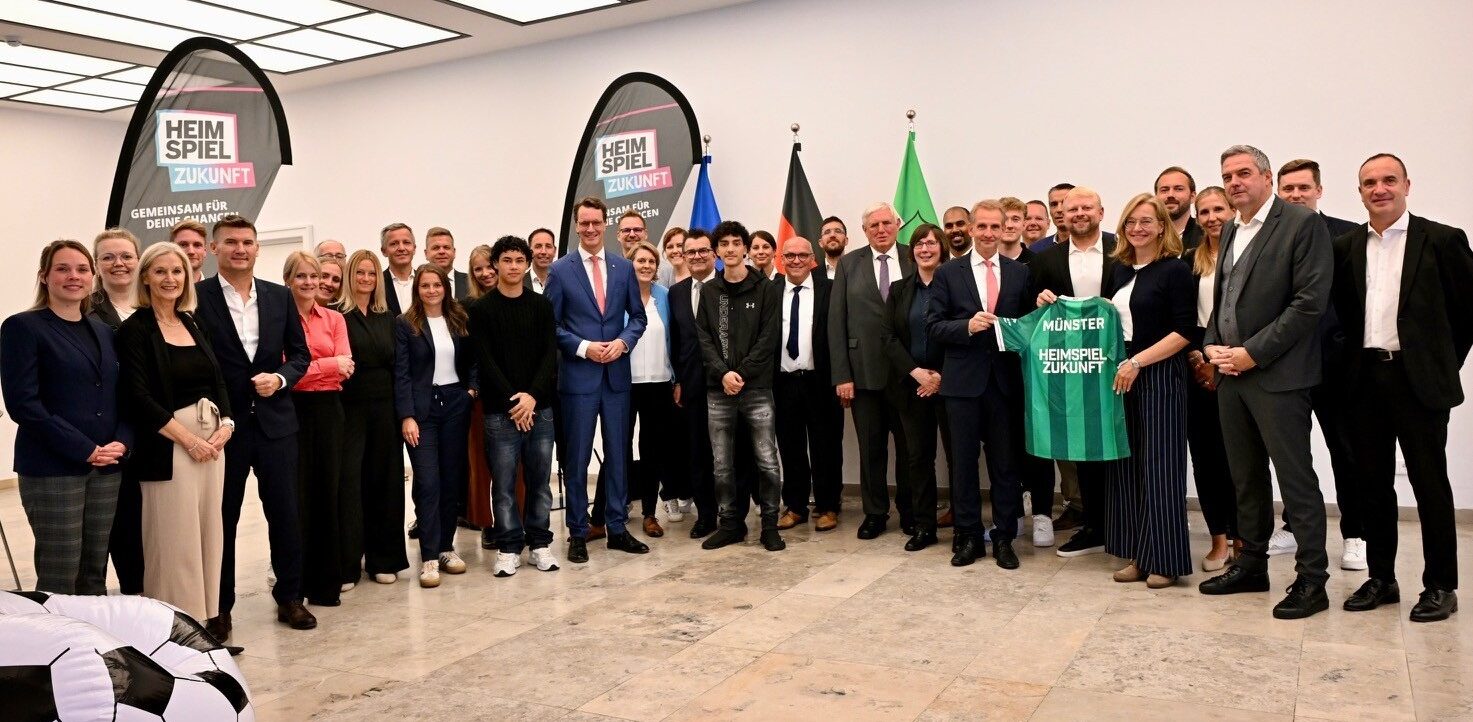Gruppenfoto einer Veranstaltung der Fußballbundnis mit Teilnehmern in einem Raum. Im Hintergrund sind zwei Banner mit dem Text "Heimspiel Zukunft" und "Gemeinsam für Deine Chance" zu sehen. Drei Flaggen, darunter die deutsche Flagge, sind im Hintergrund aufgestellt. In der Mitte halten zwei Personen ein grünes Trikot mit der Aufschrift "Münster Heimspiel Zukunft".